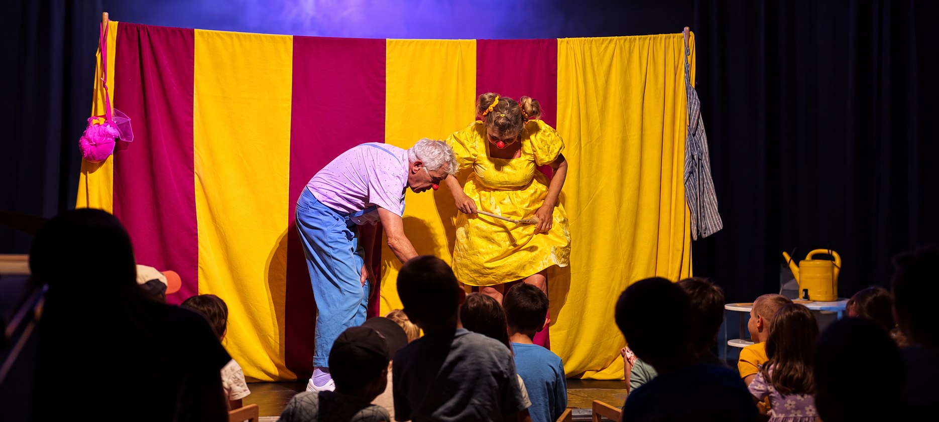 Das Clownduo spielt vor gelben und magentafarbenen Vorhängen; im Vordergrund sitzt das Publikum.