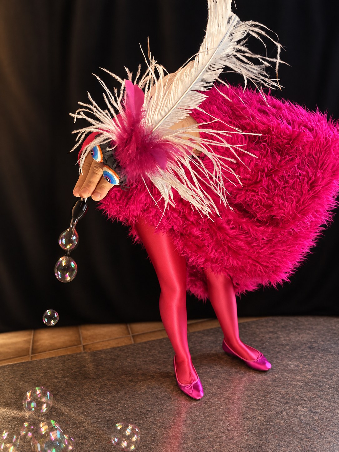 Eine zottelige rosa Vogelfigur mit langen weißen Federn pustet Seifenblasen.